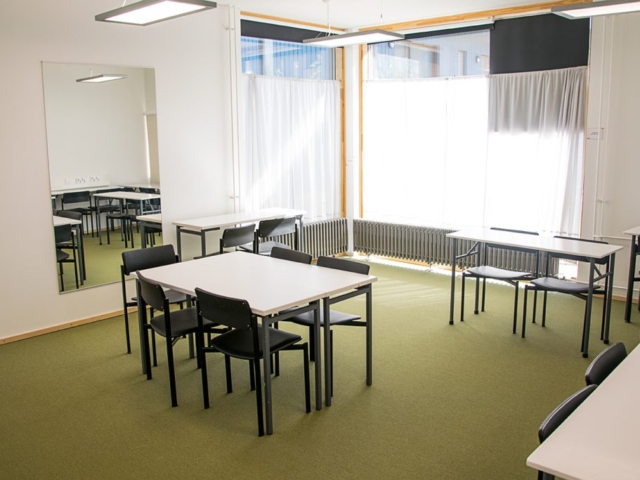 Tilan toinen puoli: nurkkauksessa ikkunoita, vasemmalla seinustalla peili, erikokoisia pöytäryhmiä.