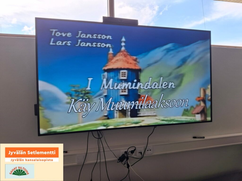 Kuva televisiosta seinällä, jossa on meneillään Muumit. Vasenmassa alakulmassa Jyvälän Setlementin logo ja slogan, sekä Sanat Siltoina -hankkeen logo.