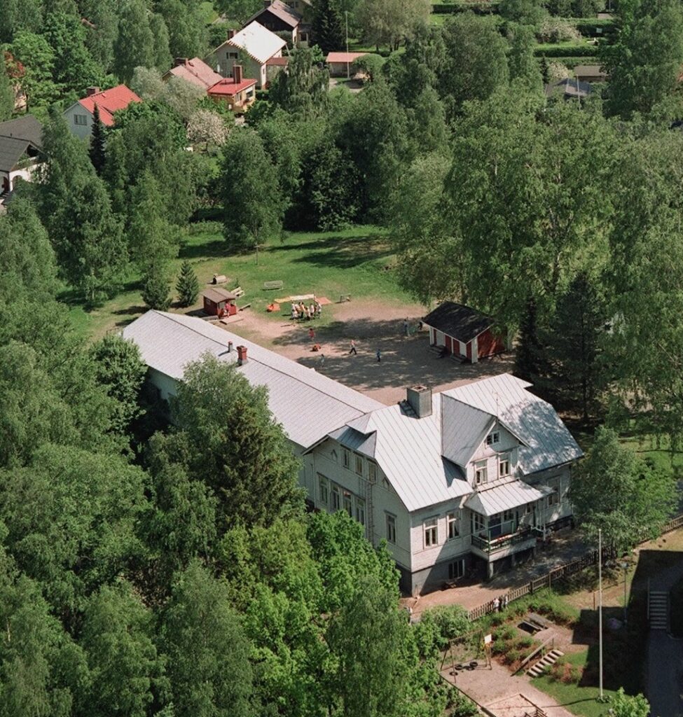 Jyvälän vanha valkoinen rakennus kuvattuna yläpuolelta puiden saartamana. Takapihalla näkyy ihmisiä pelaamassa.