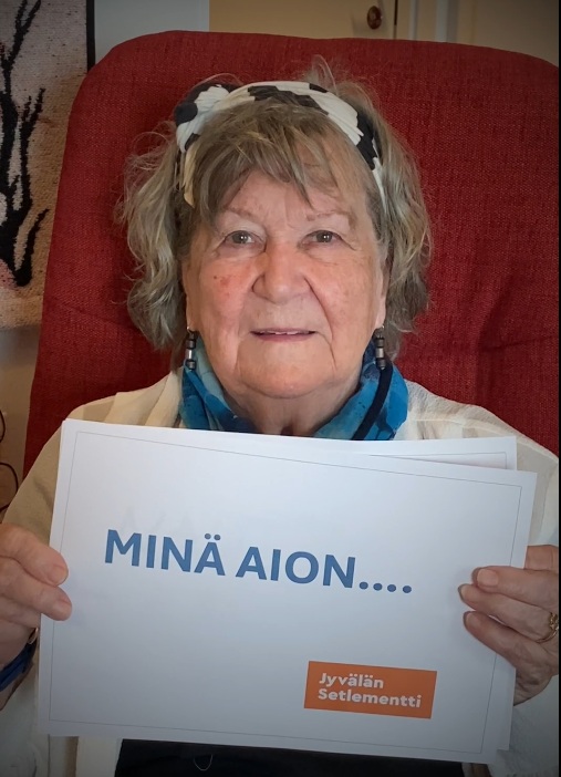 Kuva naisesta istumassa punaisessa nojatuolissa pitämässä molemmissa käsissä paperia, jossa lukee "Minä aion..." Jyvälän Setlementti logolla.
